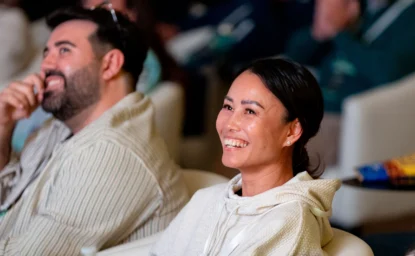 Two people sit side by side in an audience, smiling and looking towards the front of the room.
