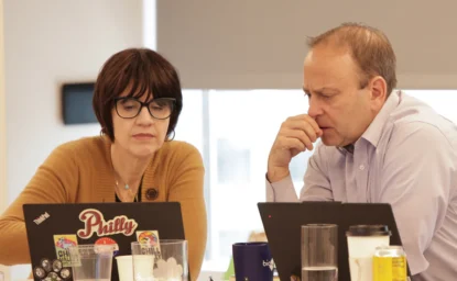 Two people sit at a table working on laptops, appearing focused; various cups, bottles, and papers are on the table.