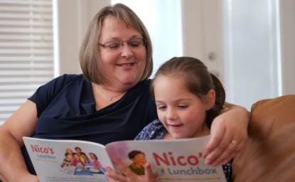 An adult and a young girl sit on a couch reading a book titled "Nico's Lunchbox" together, both smiling and engaged with the story.