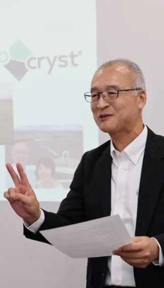 An older man in a suit holds papers and makes a peace sign gesture while standing in front of a projection screen displaying images and text.