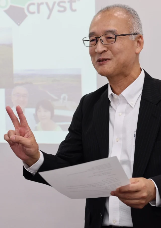 A man in a suit holds papers and gestures with a peace sign while speaking in front of a projected presentation.