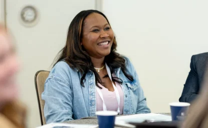 A smiling woman in a denim jacket sits at a table during a meeting.